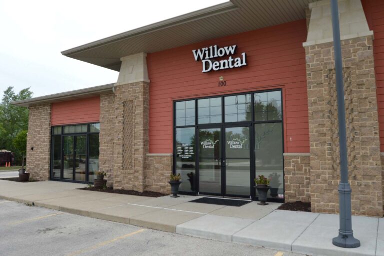 Exterior of a modern dental office named “Willow Dental,” featuring a mix of brick and red siding with glass doors and windows. Two potted plants flank the entrance, and the address “100” is displayed above. Sidewalk and parking lot visible.