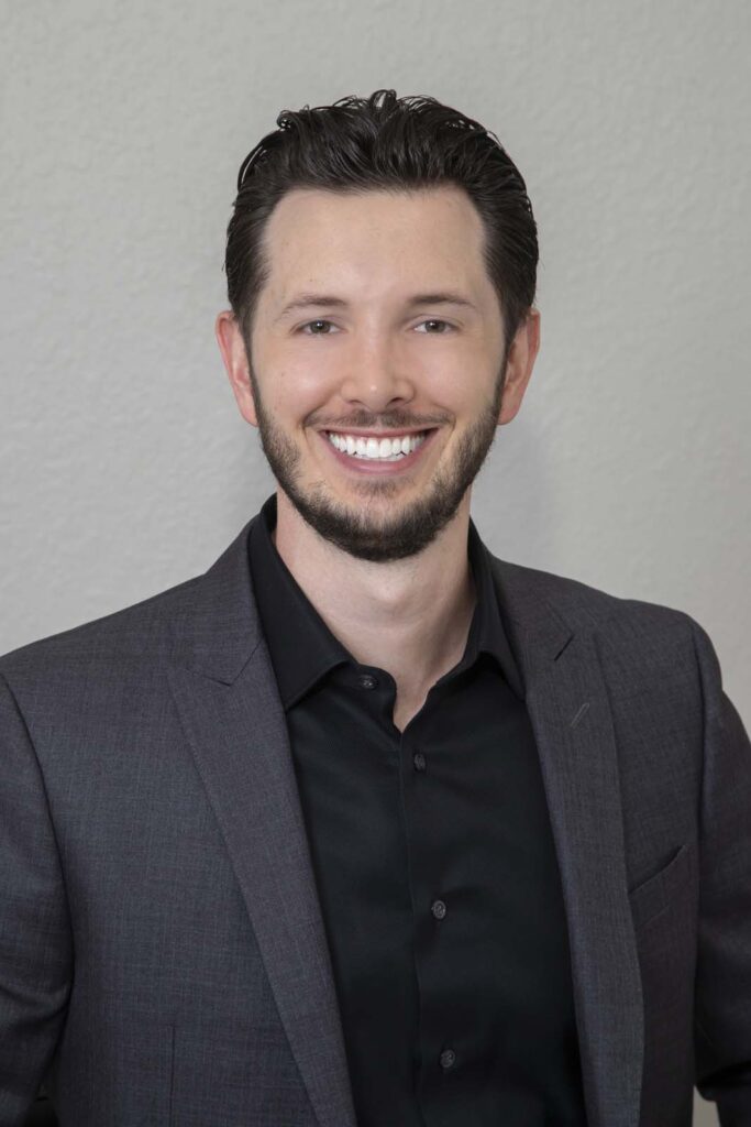 Dr. Jake Jacomet of Willow Dental in New Berlin, WI. The image shows a person wearing a dark grey suit jacket over a black dress shirt. The background is a plain light grey wall. It appears to be a professional portrait or headshot.