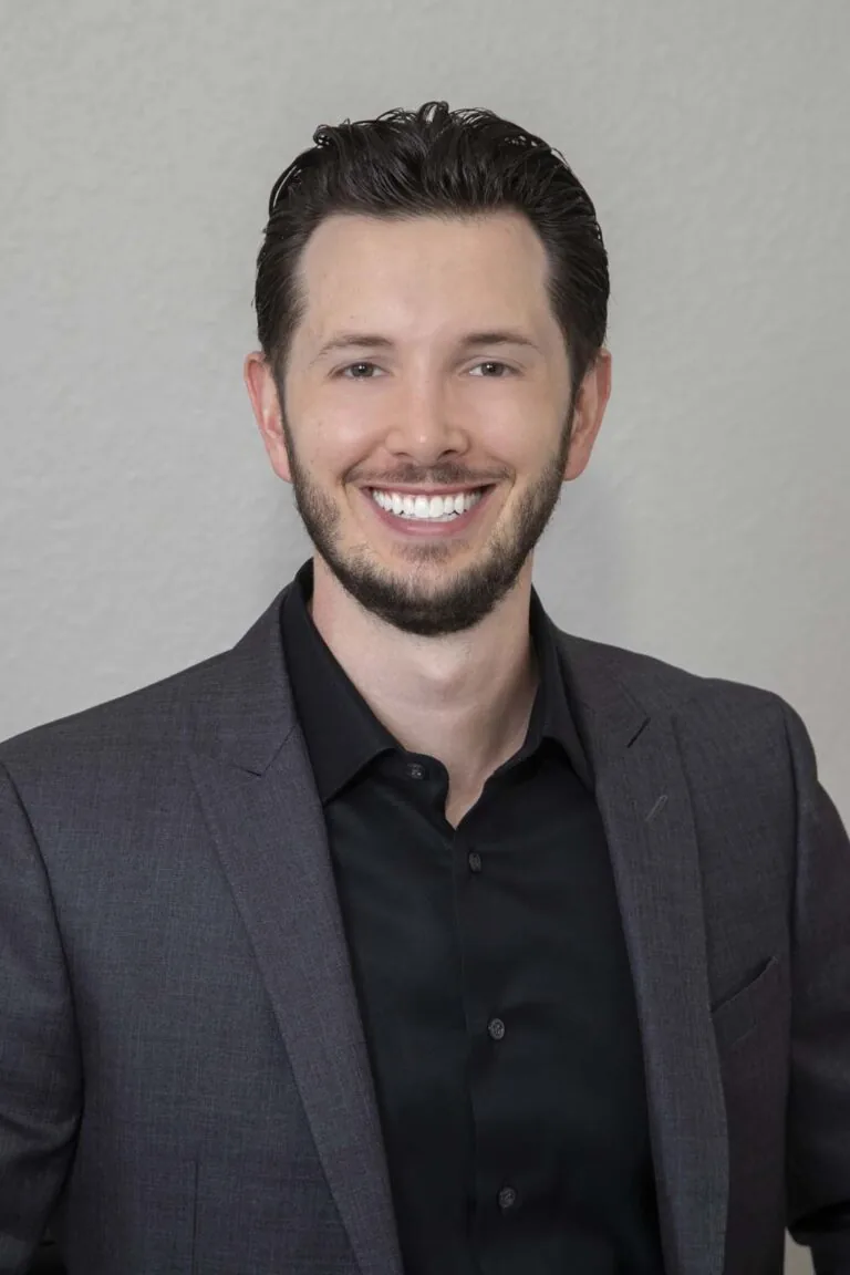 Dr. Jake Jacomet of Willow Dental in New Berlin, WI. The image shows a person wearing a dark grey suit jacket over a black dress shirt. The background is a plain light grey wall. It appears to be a professional portrait or headshot.