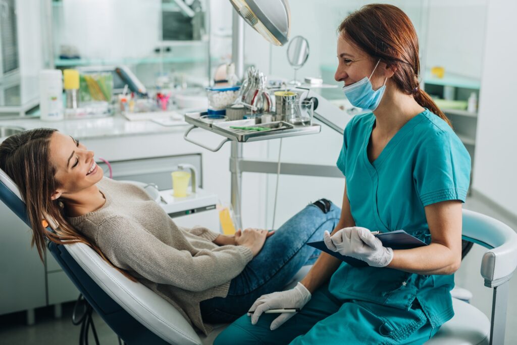 The image shows a dental clinic setting with a patient reclining in a dental chair. A dentist is seated nearby, wearing blue scrubs and gloves, and holding a clipboard.