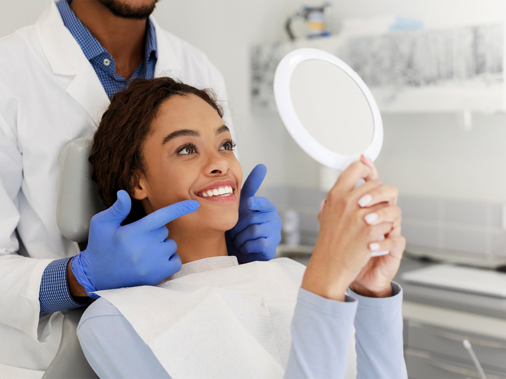 The image shows a person sitting in a dental chair, holding a round mirror while wearing a dental bib. A dentist, dressed in a white coat and blue gloves, stands behind them, seemingly assisting or guiding their examination. The background includes typical dental office equipment and cabinetry, reinforcing the clinical setting.
