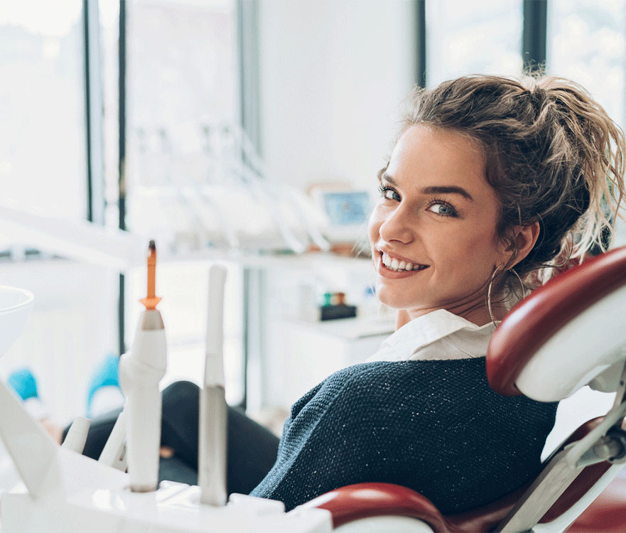A woman sitting in a dental chair in a clinical setting. The woman's face is blurred for privacy. The background includes dental equipment and tools, indicating a dental clinic environment.