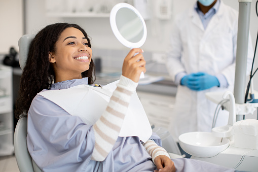 Happy Female Patient Looking At Mirror After Dental Treatment In Clinic, Cheerful American Woman Sitting In Chair In Stomatological Cabinet And Enjoying Her Beautiful Smile, Closeup